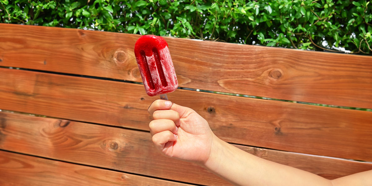 Person holding red popsicle in front of wooden bench. | Une personne tient une glace rouge devant un banc en bois.