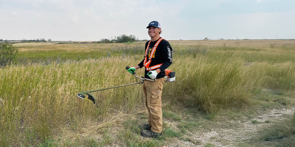 A man operates a weed trimmer in a lush green field, focused on maintaining the landscape. Un homme utilise un coupe-herbe dans un champ verdoyant et luxuriant, en se concentrant sur l'entretien du paysage.