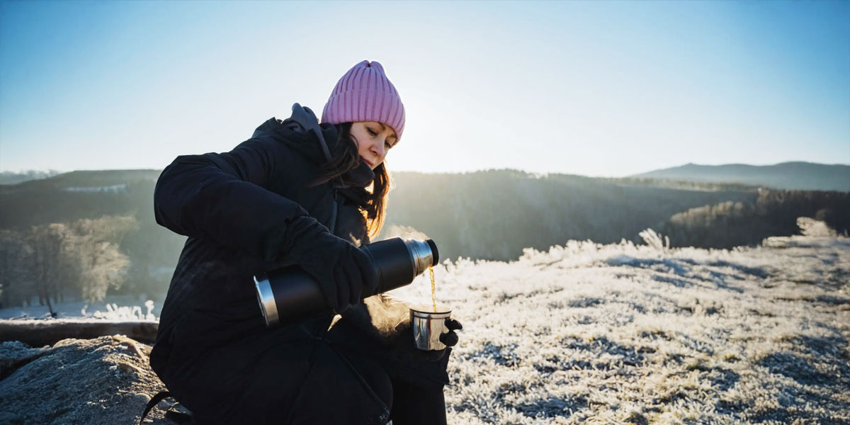 A woman wearing a pink hat crouching on a trail, enjoying a cup of tea in a serene outdoor setting. Une personne portant un chapeau rose, accroupie sur un tronçon du Sentier, savourant une tasse de thé dans un cadre naturel paisible.