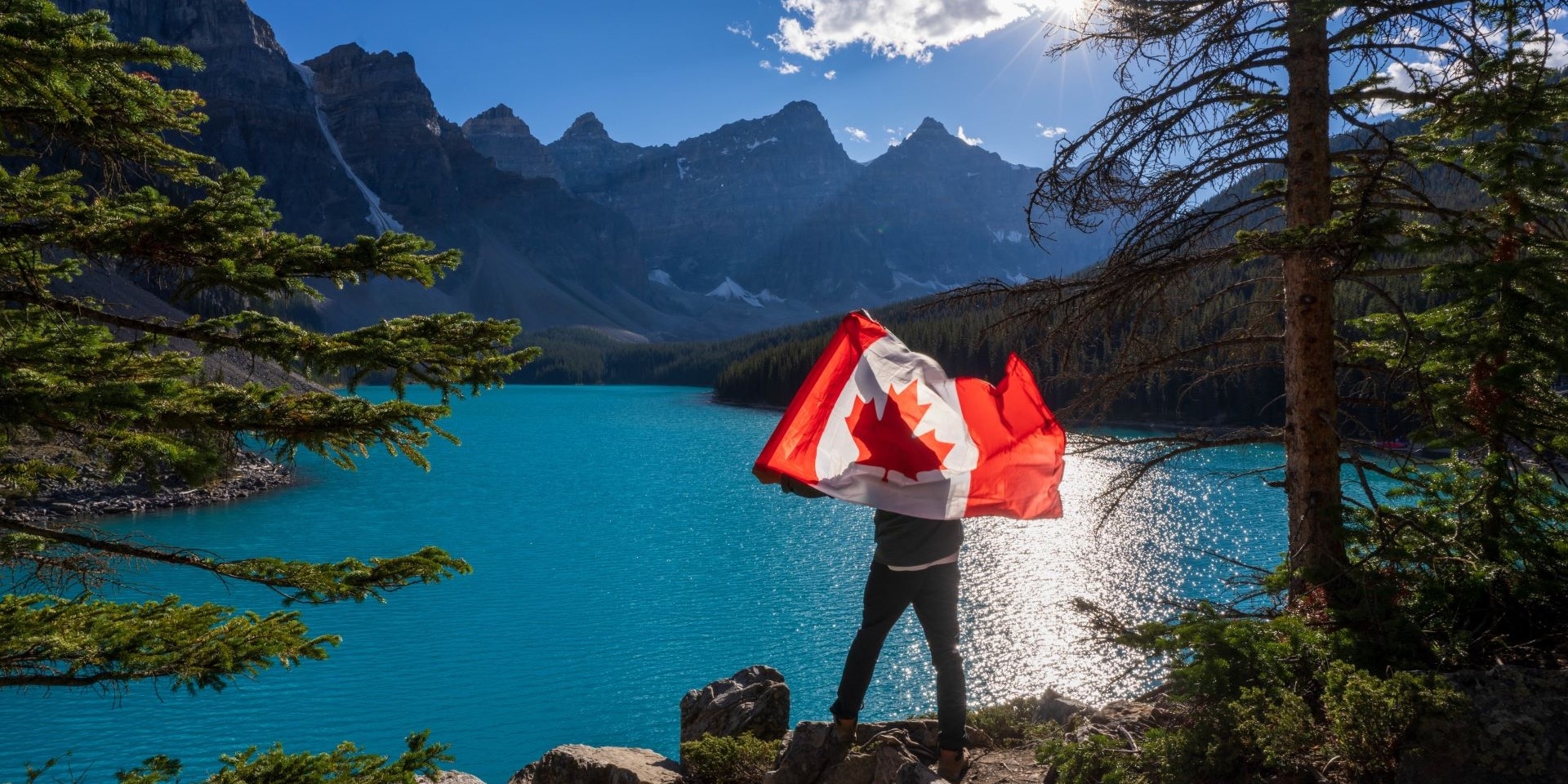 flag-mountains-tct-homepage | Trans Canada Trail A person holds a Canadian flag on a cliff above a lake with mountains in the background.