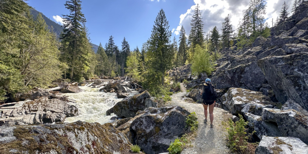 person walking along a trail with river and forest and blue skies