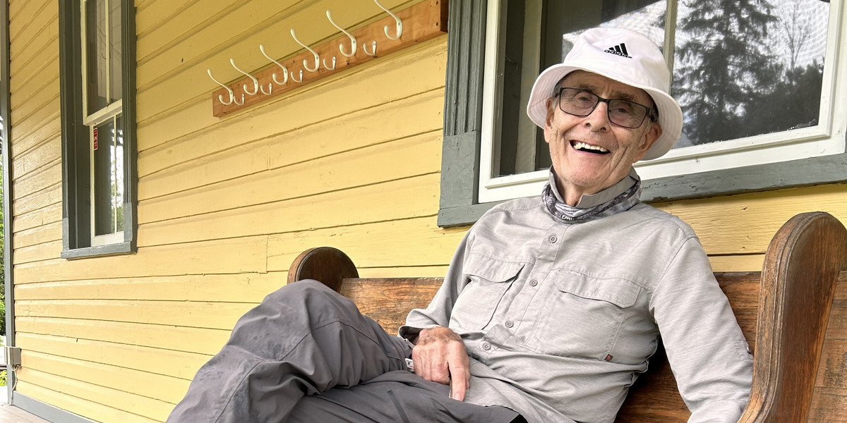 QC_Web story_Feature image_Volunteer recognition Ken Bouchard_credit Sentier Wakefield Trails | Trans Canada Trail Man sitting on bench outside a yellow house, smiling at the camera.