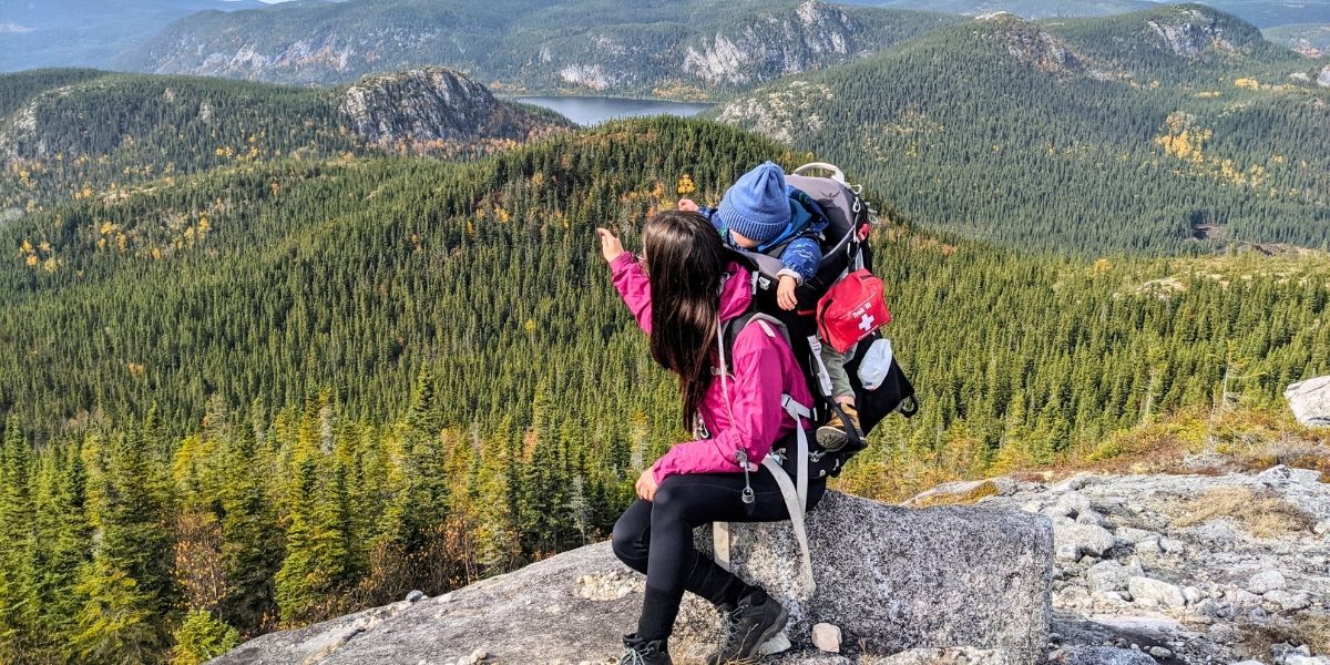 A person sitting on a rock with a baby in a backpack on their back. They are looking at the mountain and valley view of evergreen trees.