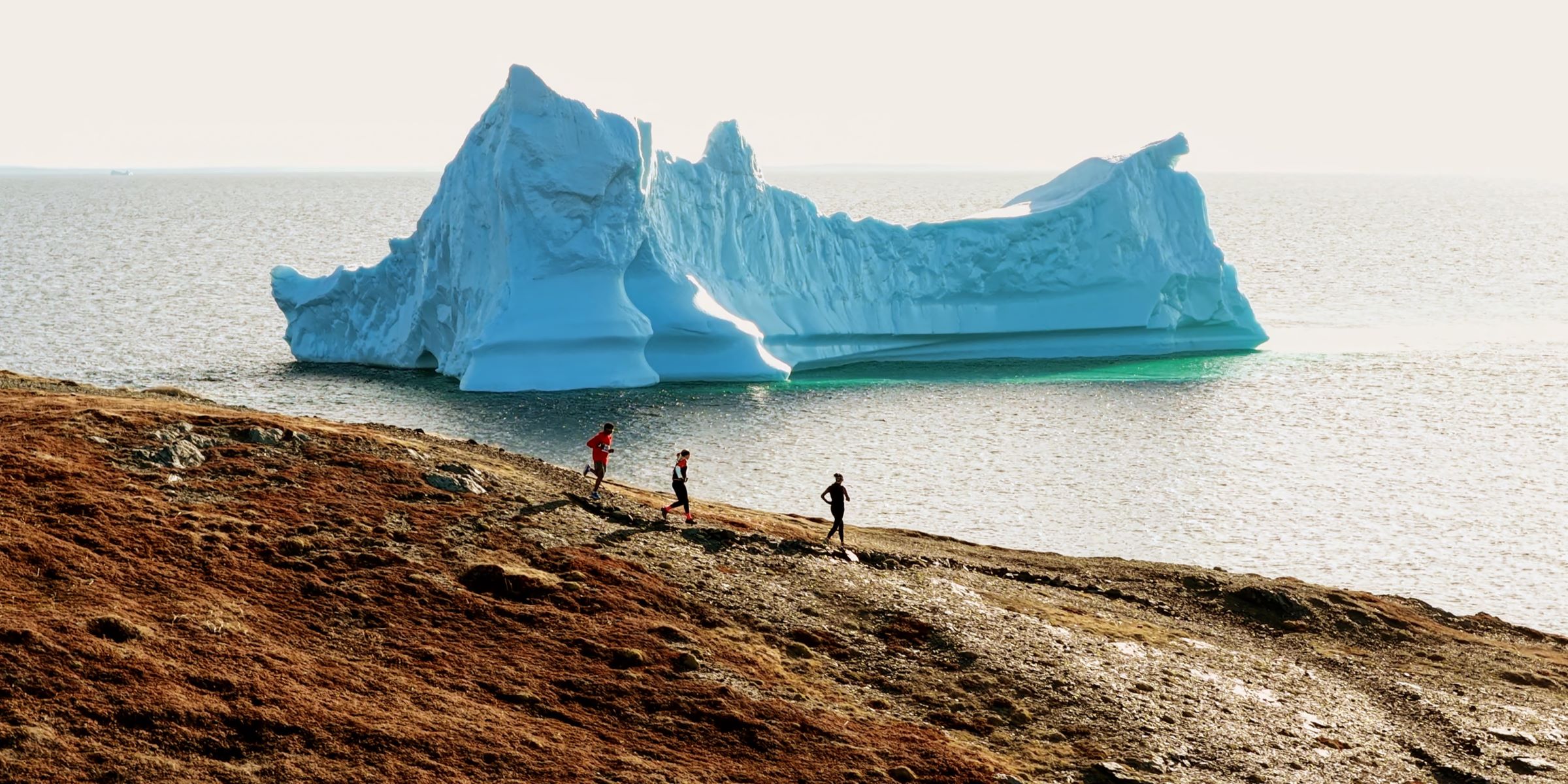 People run on the Atlantic coast along the Great Northern Trail with an iceberg in the background.