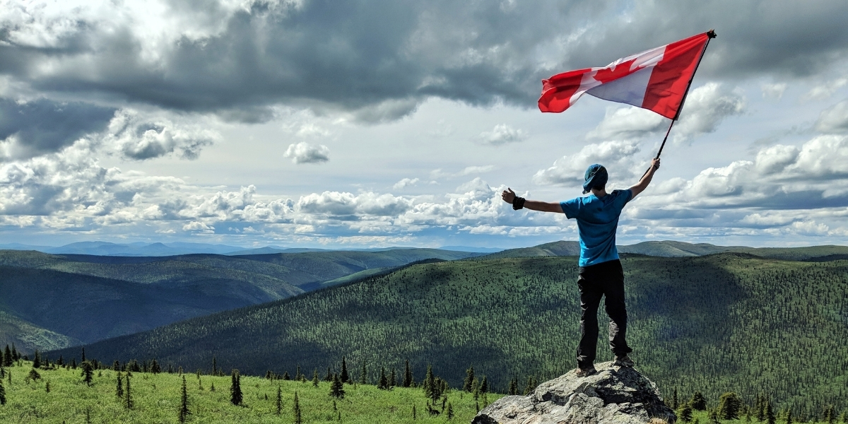 00Feature image_Aidan Beckett_credit Aidan Beckett | Trans Canada Trail man holding a canadian flag on a mountain top on a cloudy day