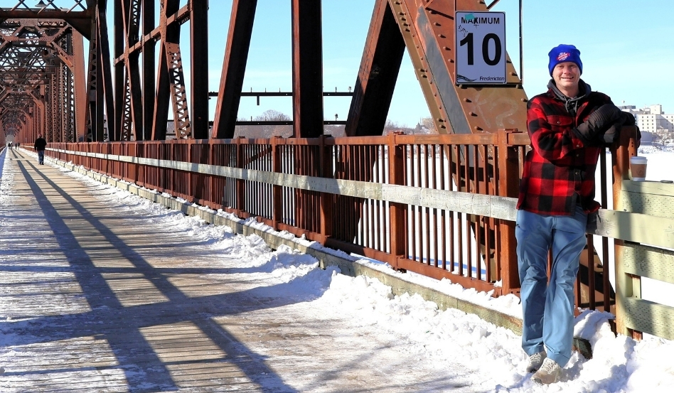 00Feature image_Cathy Donaldson_Greg McCann image_credit Cathy Donaldson | Trans Canada Trail Man in a red plaid jacket and blue beanie leaning on the railing of a snow-covered metal bridge, with a 'Maximum 10' sign nearby.