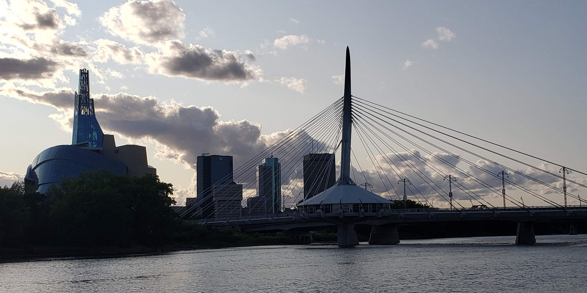Cable-stayed bridge with a tall central pylon spanning a calm river, set against a city skyline with a distinctive curved blue building on the left at dusk/light clouds above.