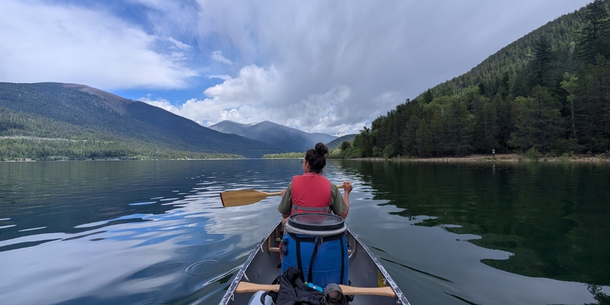Person wearing a pink life vest paddles a canoe across a calm mountain lake, with forested hills and mountains under a partly cloudy sky (back view).