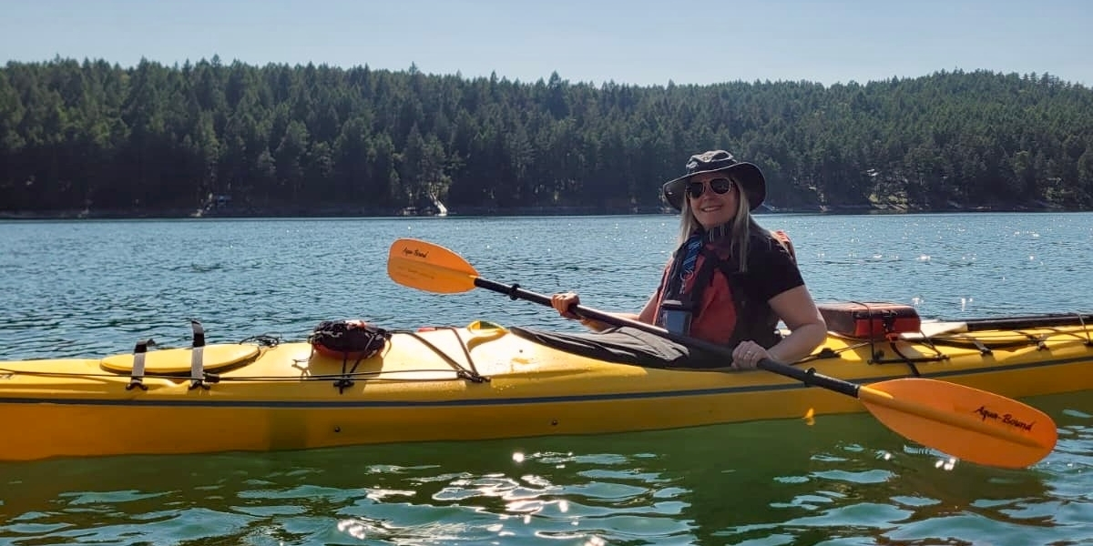 00Feature image_Claudia Laroye_Salish Sea Marine Trail_credit Claudia Laroye | Trans Canada Trail Woman wearing a wide-brim hat and sunglasses paddling a yellow kayak on a calm lake with a forested shoreline in the background, holding an orange paddle.