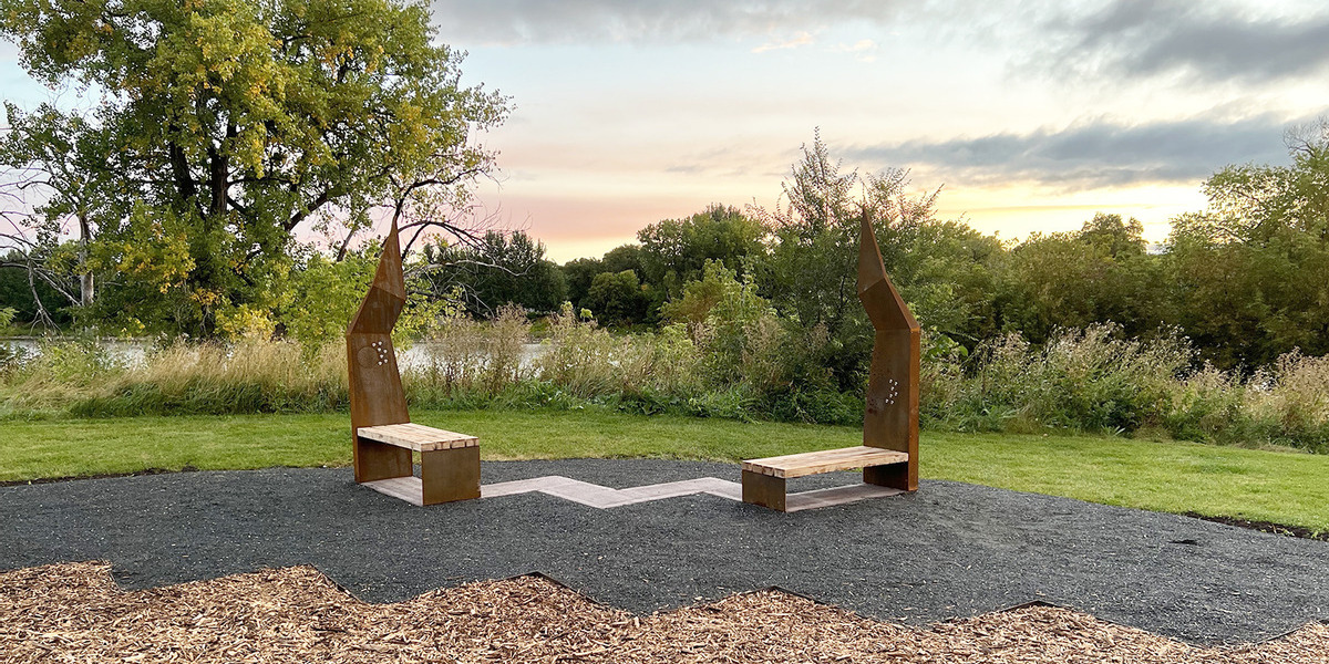 SF104196_MB_The-Speech-Act-Project_City-of-Winnipeg-3_Web_StoryFeatured_Image_1200x600 | Trans Canada Trail Two rusty angular benches face each other by a paved path in a park at sunset, near tall grasses and a lake on the horizon, artful seating.